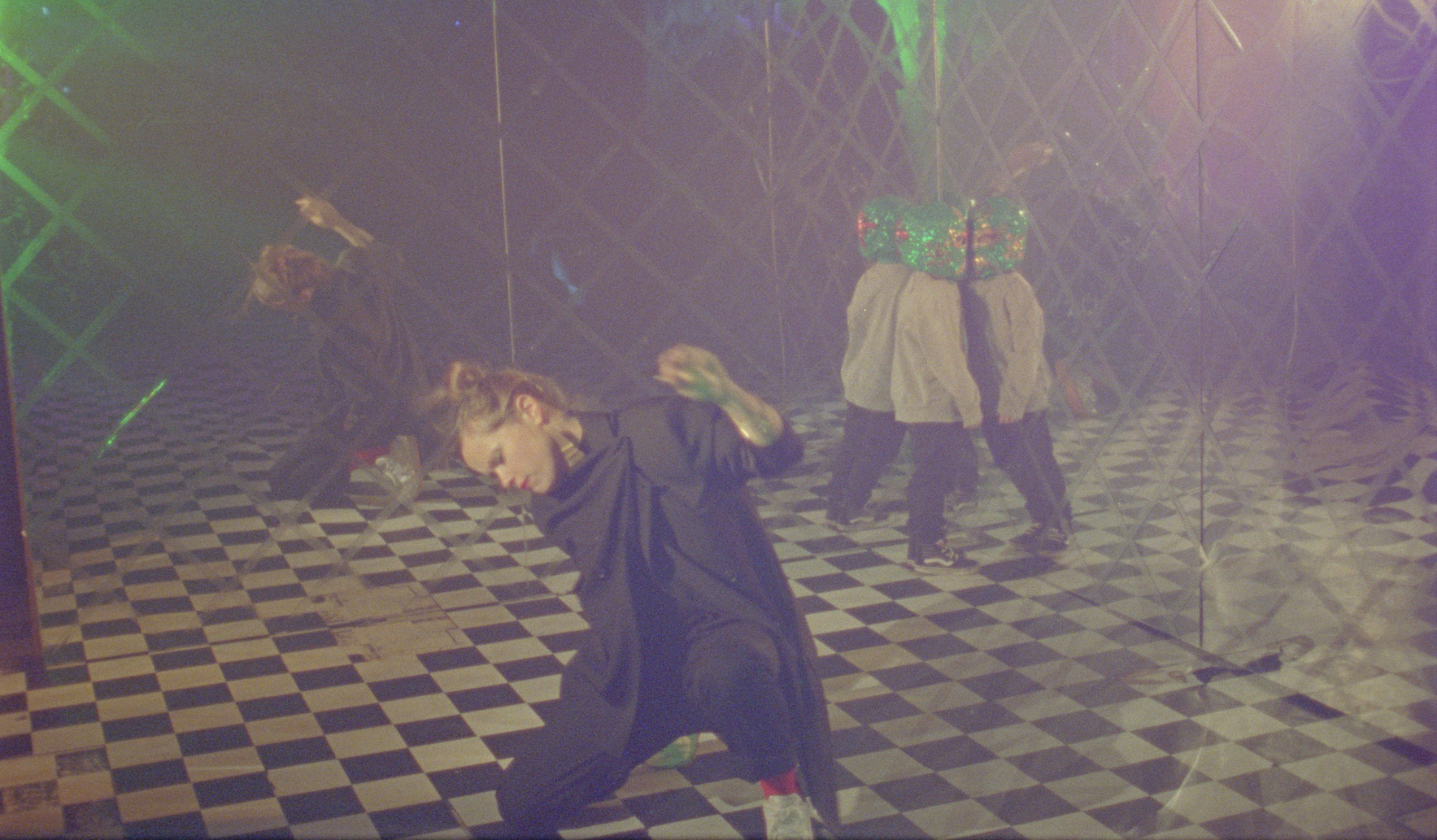 Interior shot of a nightclub with black and white floor, and mirrored walls. A white-skinned woman with blonde hair tied up in a messy top-knot appears to breakdance on the floor. She is wearing a draped black two-piece, red socks and white sneakers. She wears red lipstick and metal jewellry. In the rear of the room, a small child pushes his whole body up against the mirror wall. He is wearing an oversize green sequined Lucha libre mask, a grey sweater, black trousers and dark sneakers. A disco ball refracts blue, purple and green light all over the dancefloor.