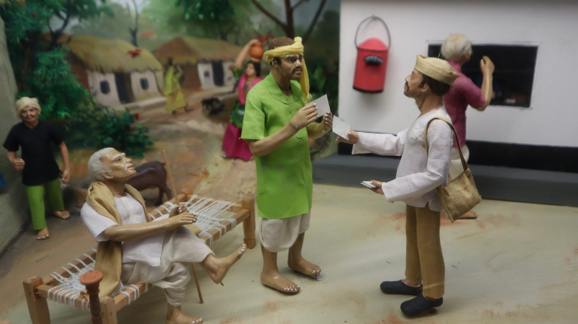 Colour image of a miniature diorama of cartoon figurines in a village scene. Two brown-skinned moustached figures in the foreground exchange letters, and a red postbox appears behind them. An older figure is seated on a bed, while in the background three other figures mill about, including a woman in a pink and green sari carries a jug on her head.