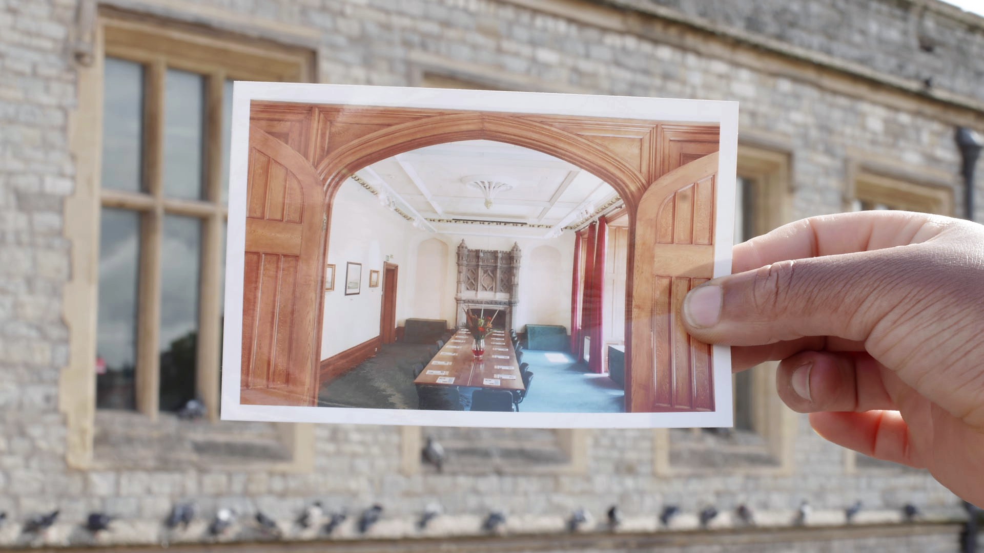 A brown-skinned hand holds out a photograph, pinched between thumb and fingers, of an old English boardroom interior filled with a line of chairs, a long table, a flower arrangement and a medieval-looking stone fireplace. In the background of the shot, behind the hand holding the photograph, is the exterior of an old building that seems to be the same sandy stone colour as the boardroom interior. Blurry and out of focus, a line of pigeons visibly rest on the bottom of the building.