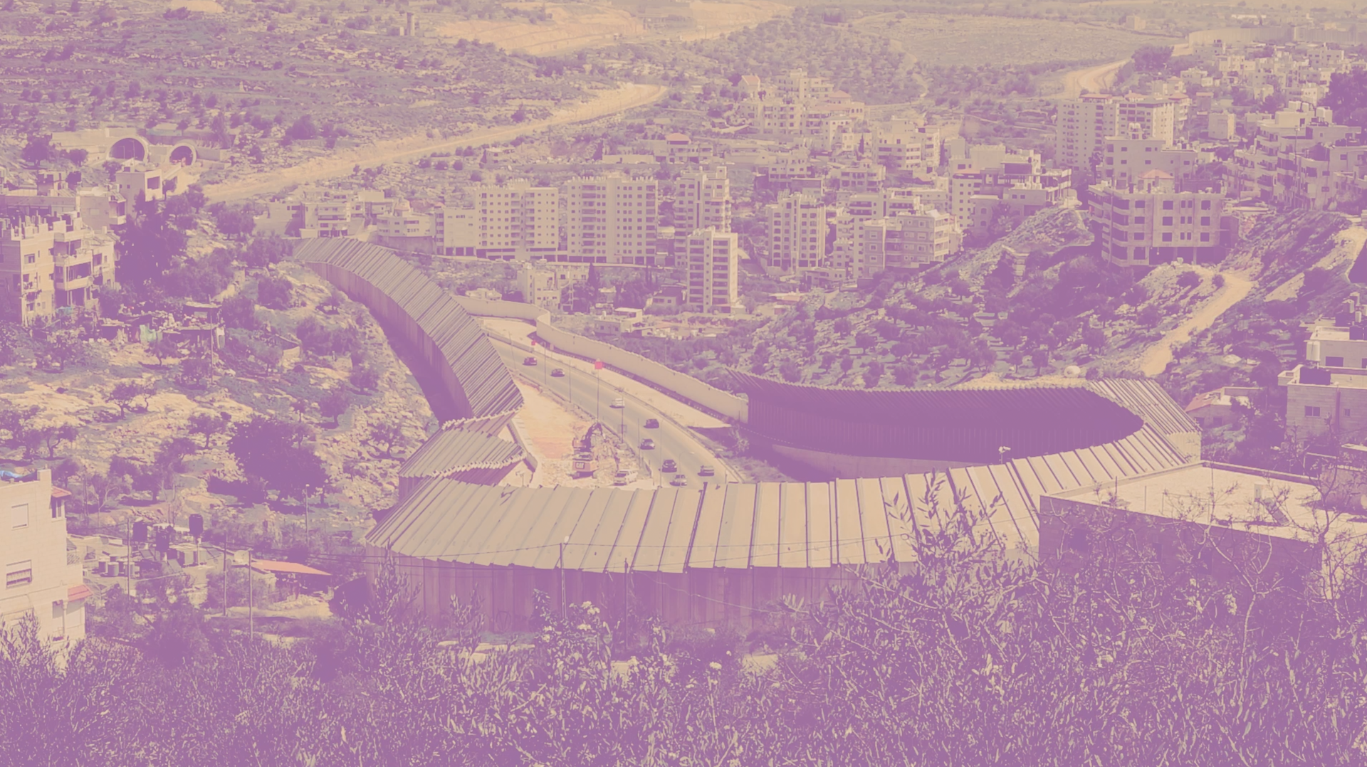 Landscape image of a snaking highway enclosed by high walls that shield the view of the landscape. Built up condos appear on the right, with olive trees in the foreground.